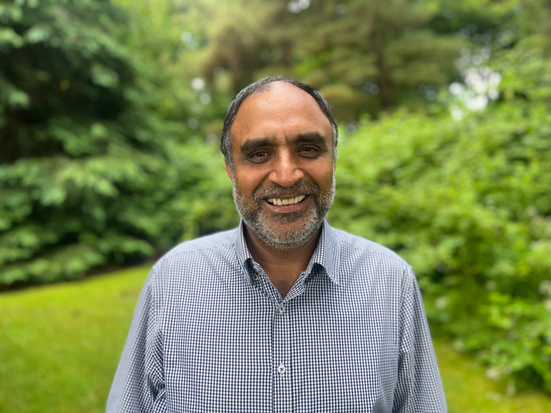 A man with a beard, wearing a blue checked shirt, smiles warmly while standing outdoors.